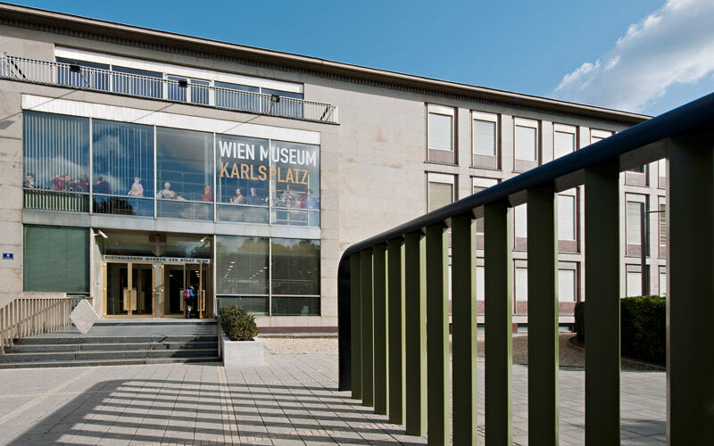 Außenansicht des Wien Museum Karlsplatz mit großen Glasfenstern, Eingangstreppe und grünem Geländer im Vordergrund an einem klaren Tag.