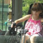 Ein junges Mädchen in einem rosafarbenen Kleid sitzt in einem Baufahrzeug, hält die Steuerhebel in der Hand und blickt nach vorne.