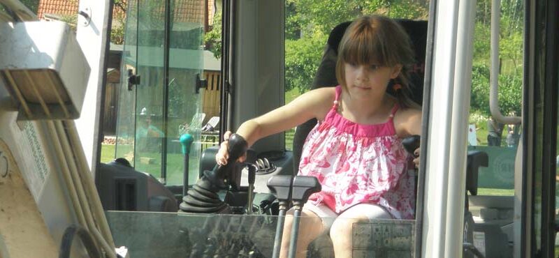 Ein junges Mädchen in einem rosafarbenen Kleid sitzt in einem Baufahrzeug, hält die Steuerhebel in der Hand und blickt nach vorne.