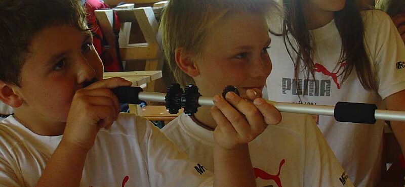 Zwei Jungen in weißen T-Shirts halten und untersuchen einen Metallstab mit schwarzen Griffen, umgeben von anderen Kindern in einem Haus.