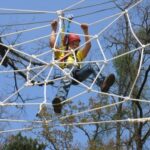 Ein Kind, das einen Helm und einen Klettergurt trägt, klettert im Freien, umgeben von Bäumen und einem strahlend blauen Himmel, auf eine Seilnetzstruktur.
