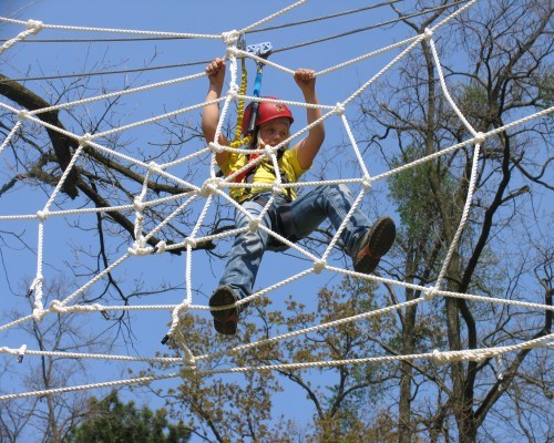 Ein Kind, das einen Helm und einen Klettergurt trägt, klettert im Freien, umgeben von Bäumen und einem strahlend blauen Himmel, auf eine Seilnetzstruktur.