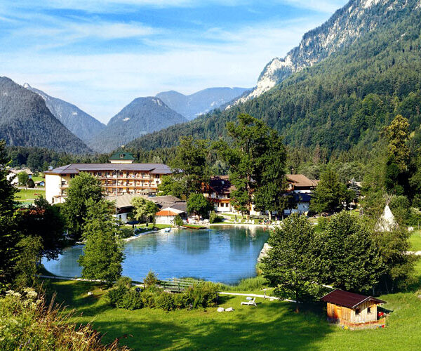 Ein kleiner See, umgeben von Bäumen, Gebäuden und Bergen unter blauem Himmel; im Vordergrund steht eine Holzhütte.