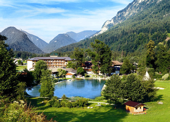 Ein kleiner See, umgeben von Bäumen, Gebäuden und Bergen unter blauem Himmel; im Vordergrund steht eine Holzhütte.