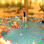 Mehrere Kinder und Erwachsene spielen in einem mit bunten Plastikbällen gefüllten Hallenbad, an dessen Wand im Hintergrund ein Wandgemälde mit Tieren und Natur zu sehen ist.