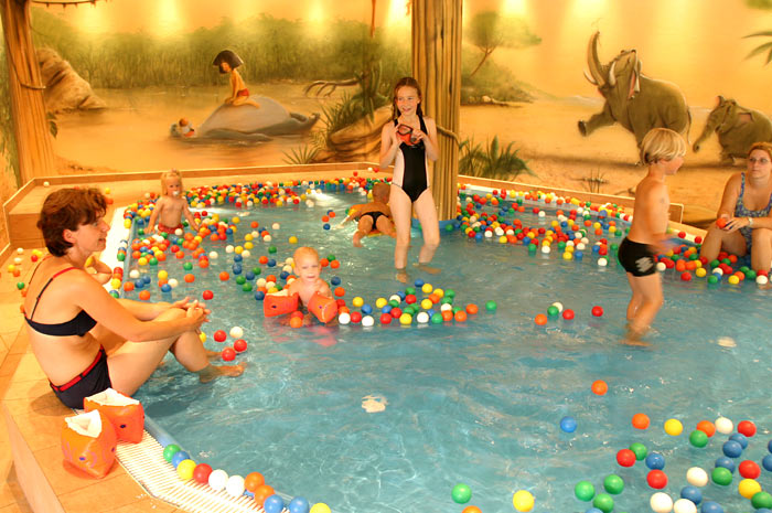 Mehrere Kinder und Erwachsene spielen in einem mit bunten Plastikbällen gefüllten Hallenbad, an dessen Wand im Hintergrund ein Wandgemälde mit Tieren und Natur zu sehen ist.