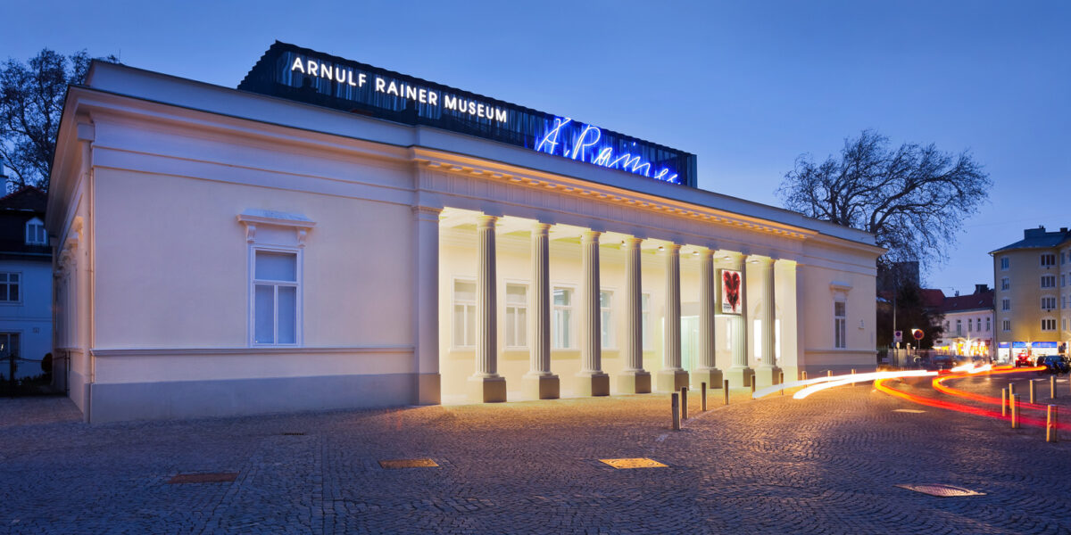 Ein neoklassizistisches Gebäude mit Säulen in der Dämmerung, beleuchtet von Lichtern, mit der Aufschrift "Arnulf Rainer Museum" und Leuchtreklame auf dem Dach. Im Vordergrund sind Lichtspuren von vorbeifahrenden Fahrzeugen zu sehen.