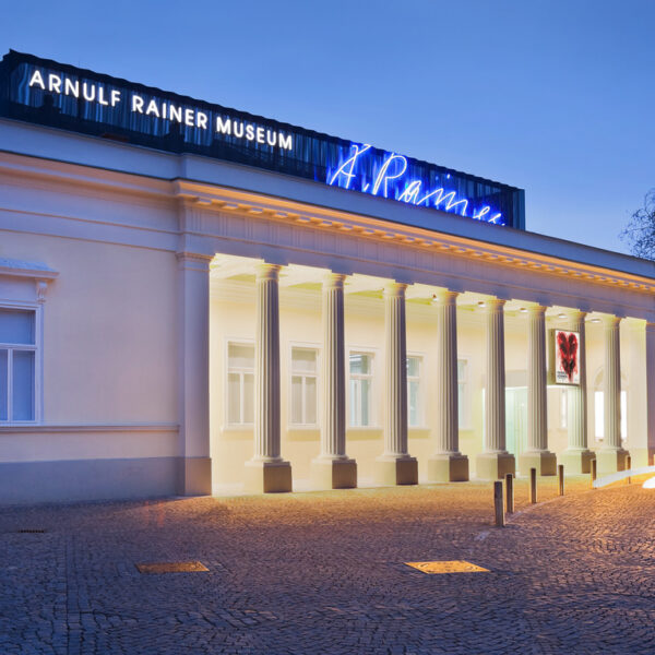 Ein neoklassizistisches Gebäude mit Säulen in der Dämmerung, beleuchtet von Lichtern, mit der Aufschrift "Arnulf Rainer Museum" und Leuchtreklame auf dem Dach. Im Vordergrund sind Lichtspuren von vorbeifahrenden Fahrzeugen zu sehen.