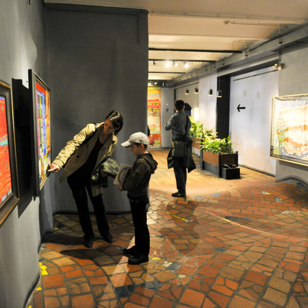 Eine Frau und ein Kind betrachten ein gerahmtes Kunstwerk an einer Galeriewand, während zwei Personen im Hintergrund andere Kunstwerke betrachten.