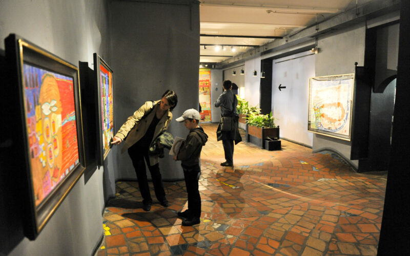 Eine Frau und ein Kind betrachten ein gerahmtes Kunstwerk an einer Galeriewand, während zwei Personen im Hintergrund andere Kunstwerke betrachten.