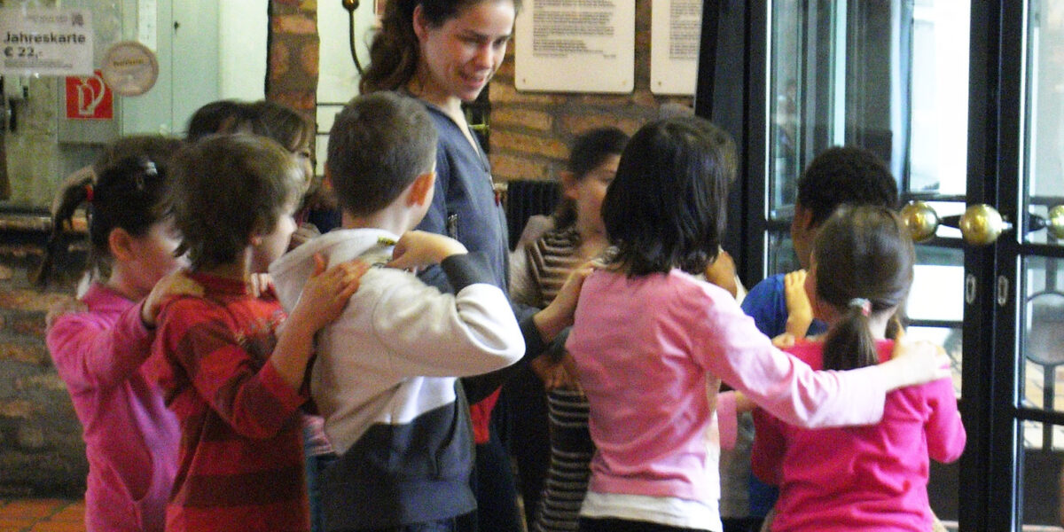 Eine Gruppe von Kindern steht im Kreis, jedes Kind hat die Hände auf den Schultern des Kindes vor ihm, während eine erwachsene Frau in der Nähe in einem Gebäude steht.