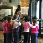 Eine Gruppe von Kindern steht im Kreis, jedes Kind hat die Hände auf den Schultern des Kindes vor ihm, während eine erwachsene Frau in der Nähe in einem Gebäude steht.