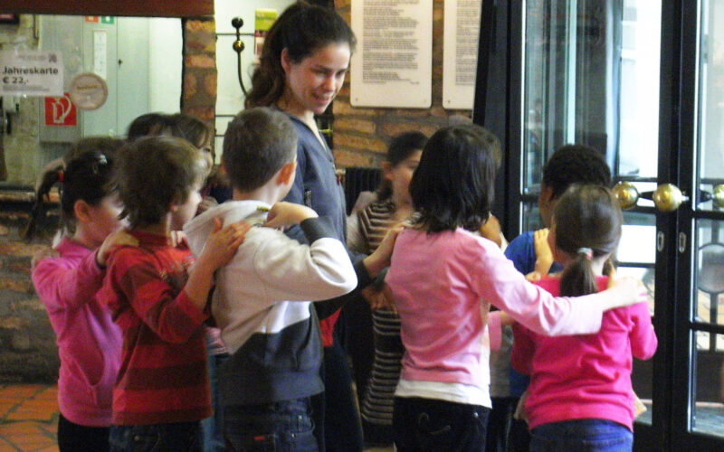 Eine Gruppe von Kindern steht im Kreis, jedes Kind hat die Hände auf den Schultern des Kindes vor ihm, während eine erwachsene Frau in der Nähe in einem Gebäude steht.