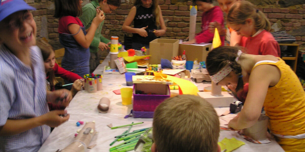 Eine Gruppe von Kindern versammelt sich um einen mit Bastelmaterial bedeckten Tisch und arbeitet in einem Raum mit Backsteinwänden an verschiedenen Kunstprojekten.