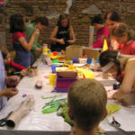 Eine Gruppe von Kindern versammelt sich um einen mit Bastelmaterial bedeckten Tisch und arbeitet in einem Raum mit Backsteinwänden an verschiedenen Kunstprojekten.