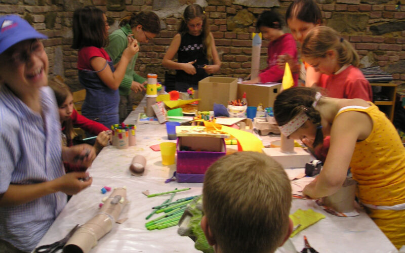 Eine Gruppe von Kindern versammelt sich um einen mit Bastelmaterial bedeckten Tisch und arbeitet in einem Raum mit Backsteinwänden an verschiedenen Kunstprojekten.