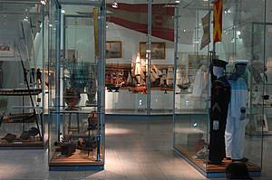 In den Glasvitrinen einer Museumsausstellung sind Marineuniformen, Hüte und Schiffsmodelle ausgestellt, während im Hintergrund verschiedene maritime Artefakte zu sehen sind.