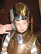 Ein kleines Kind, das eine glänzende mittelalterliche Rüstung und einen Helm trägt, während eine erwachsene Hand den Helm zurechtrückt.