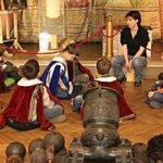 Eine Gruppe von Kindern in Kostümen sitzt auf einem Holzboden in einem Museum und hört zwei Erwachsenen zu, die vor Gemälden und einer historischen Kanone stehen.