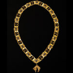 Eine goldene Zeremonienkette mit sich wiederholenden, verzierten Gliedern und einem dekorativen Anhänger auf schwarzem Hintergrund.