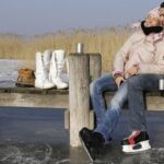 Ein Paar in Winterkleidung sitzt auf einem hölzernen Steg über einem zugefrorenen See, mit Schlittschuhen und Bechern in der Hand, mit Stiefeln und Thermoskanne in der Nähe, Schilf im Hintergrund.