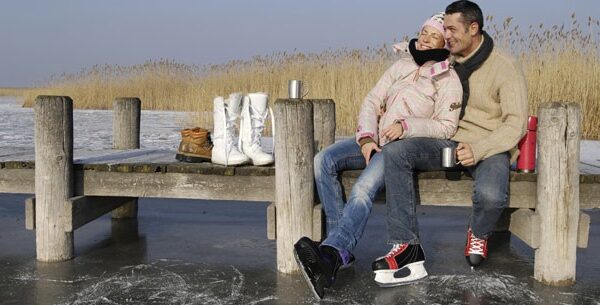 Ein Paar in Winterkleidung sitzt auf einem hölzernen Steg über einem zugefrorenen See, mit Schlittschuhen und Bechern in der Hand, mit Stiefeln und Thermoskanne in der Nähe, Schilf im Hintergrund.
