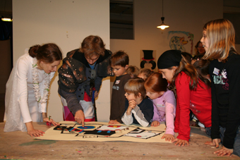 Eine Gruppe von Kindern und zwei Erwachsenen versammelt sich um einen Tisch, betrachtet und diskutiert ein buntes Kunstwerk, das vor ihnen ausgebreitet ist.