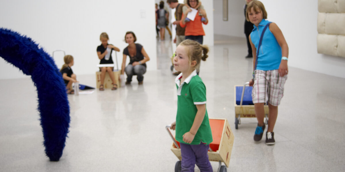 Kinder ziehen Holzkarren in einem hellen, modernen Galerieraum mit Erwachsenen und anderen Kindern im Hintergrund. Die Kunstwerke sind im ganzen Raum zu sehen.