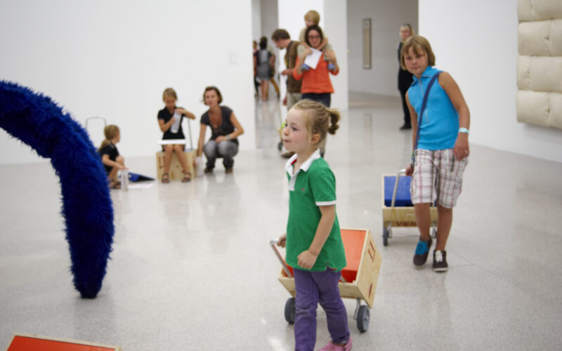 Kinder ziehen Holzkarren in einem hellen, modernen Galerieraum mit Erwachsenen und anderen Kindern im Hintergrund. Die Kunstwerke sind im ganzen Raum zu sehen.