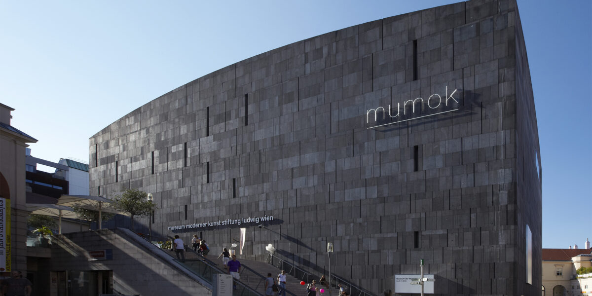 Ein großes, modernes, graues Steingebäude mit "mumok"-Schild; Menschen, die in der Nähe spazieren gehen und auf den Treppen am Eingang sitzen.