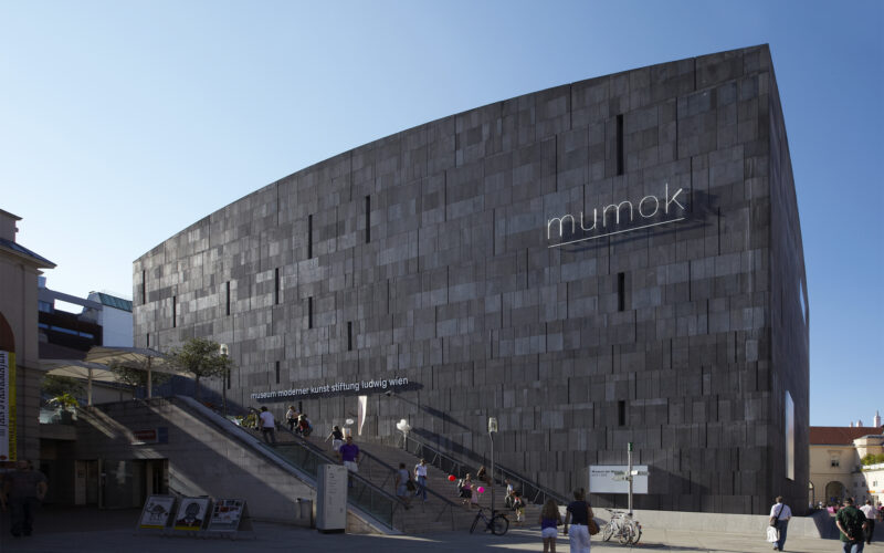Ein großes, modernes, graues Steingebäude mit "mumok"-Schild; Menschen, die in der Nähe spazieren gehen und auf den Treppen am Eingang sitzen.