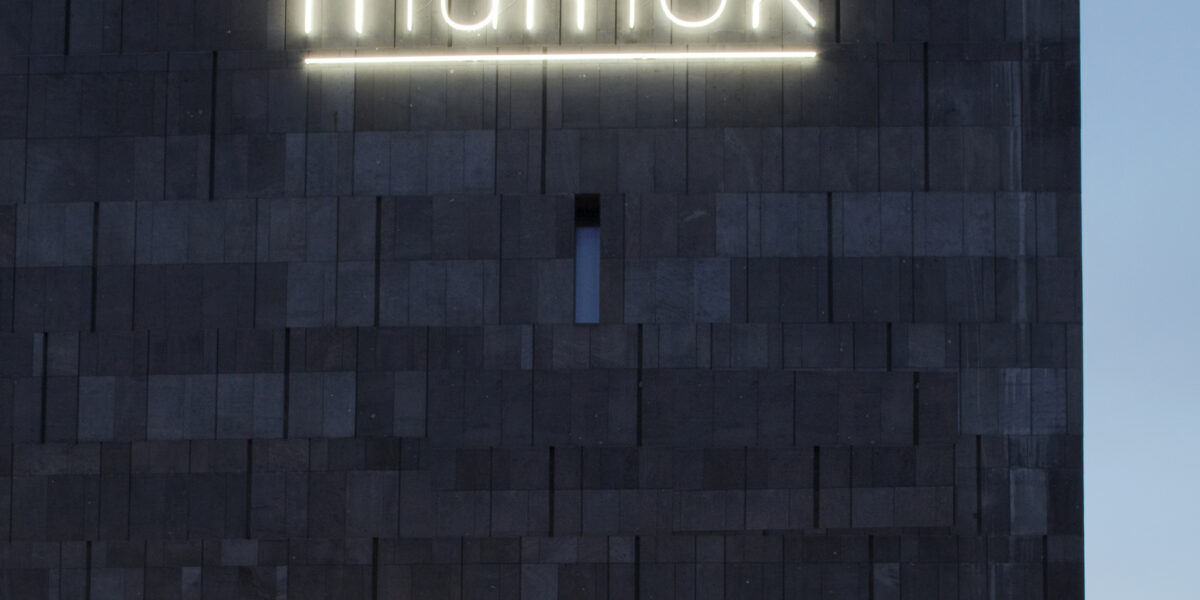 Ein dunkles Steingebäude mit beleuchtetem "mumok"-Schild, ein Museumseingang, draußen geparkte Fahrräder und eine leuchtende Straßenlaterne in der Abenddämmerung.