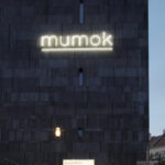 Ein dunkles Steingebäude mit beleuchtetem "mumok"-Schild, ein Museumseingang, draußen geparkte Fahrräder und eine leuchtende Straßenlaterne in der Abenddämmerung.