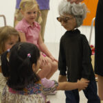 Eine Gruppe kleiner Kinder sitzt auf dem Boden und interagiert mit einer grauhaarigen Marionette, die von einem Erwachsenen in einer Galerie oder einem Klassenzimmer gesteuert wird.