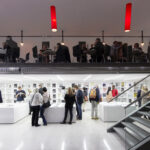 Zwei Etagen einer modernen Bibliothek oder eines Buchladens; unten schmökern Menschen in Büchern, während andere an Tischen im oberen Stockwerk sitzen und lesen. Darüber hängen rote Deckenleuchten.