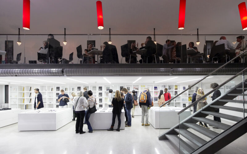Zwei Etagen einer modernen Bibliothek oder eines Buchladens; unten schmökern Menschen in Büchern, während andere an Tischen im oberen Stockwerk sitzen und lesen. Darüber hängen rote Deckenleuchten.
