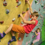Zwei Kinder klettern an einer Boulderwand in einer Halle, wobei das ältere Kind im Vordergrund und das jüngere Kind dahinter steht.