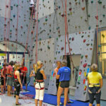 In einer Kletterhalle versammeln sich Menschen mit Klettergurten, eine Person klettert an der Wand, andere warten oder bereiten sich auf das Klettern vor.