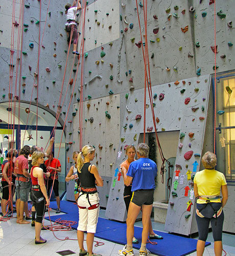 In einer Kletterhalle versammeln sich Menschen mit Klettergurten, eine Person klettert an der Wand, andere warten oder bereiten sich auf das Klettern vor.