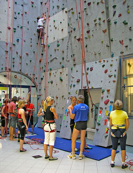 In einer Kletterhalle versammeln sich Menschen mit Klettergurten, eine Person klettert an der Wand, andere warten oder bereiten sich auf das Klettern vor.