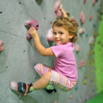 Ein kleines Kind in einem rosa Hemd und einer gestreiften Hose klettert an einer Indoor-Kletterwand und hält sich an bunten Griffen fest.