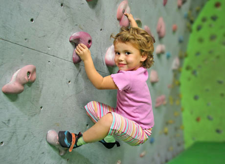 Ein kleines Kind in einem rosa Hemd und einer gestreiften Hose klettert an einer Indoor-Kletterwand und hält sich an bunten Griffen fest.