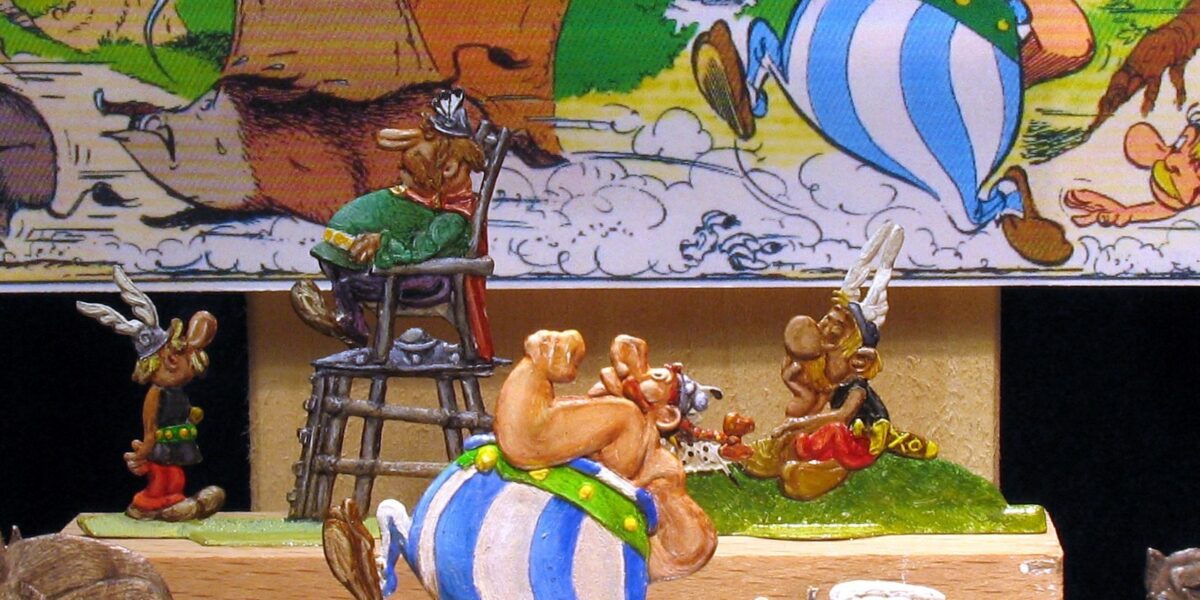 Diorama der Asterix-Comicfiguren mit Obelix, der einen Mann auf einem Stuhl trägt, vor dem Hintergrund einer Comictafel.