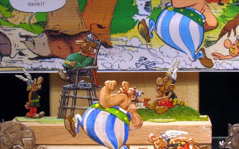 Diorama der Asterix-Comicfiguren mit Obelix, der einen Mann auf einem Stuhl trägt, vor dem Hintergrund einer Comictafel.