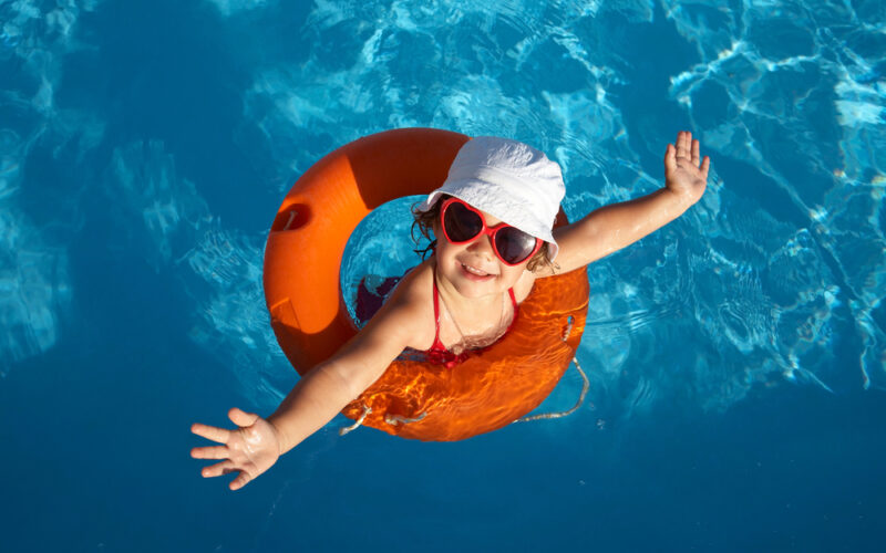 Ein Kind, das eine rote Sonnenbrille und einen weißen Hut trägt, lächelt, während es mit einem orangefarbenen Schwimmring in einem Pool schwimmt.