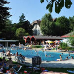 An einem sonnigen Tag entspannt man sich in einem öffentlichen Freibad, das von Gras, Bäumen, Liegestühlen und kleinen Gebäuden umgeben ist.