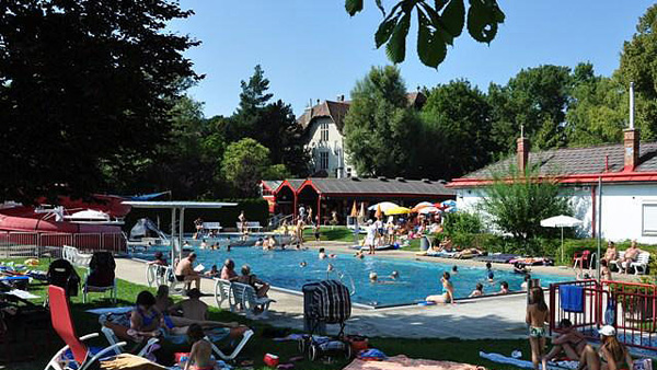 An einem sonnigen Tag entspannt man sich in einem öffentlichen Freibad, das von Gras, Bäumen, Liegestühlen und kleinen Gebäuden umgeben ist.
