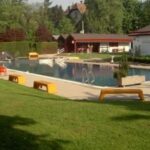 Ein Freibadbereich mit roten Wasserrutschen, Badeleitern, Bänken, grünem Gras und umliegenden Gebäuden an einem sonnigen Tag.