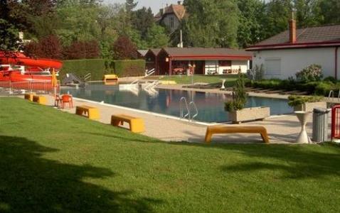 Ein Freibadbereich mit roten Wasserrutschen, Badeleitern, Bänken, grünem Gras und umliegenden Gebäuden an einem sonnigen Tag.