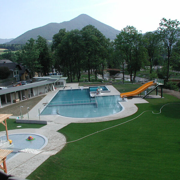 Freibadkomplex mit gelber Rutsche, grünem Rasen, umliegenden Bäumen und Bergen im Hintergrund unter einem klaren Himmel.